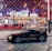 A sleek black sports car is parked in front of an illuminated arena entrance, with a large sign bearing the name of a famous beverage brand. The building features a modern, geometric facade with bright red and blue lights, creating a vibrant and lively atmosphere.